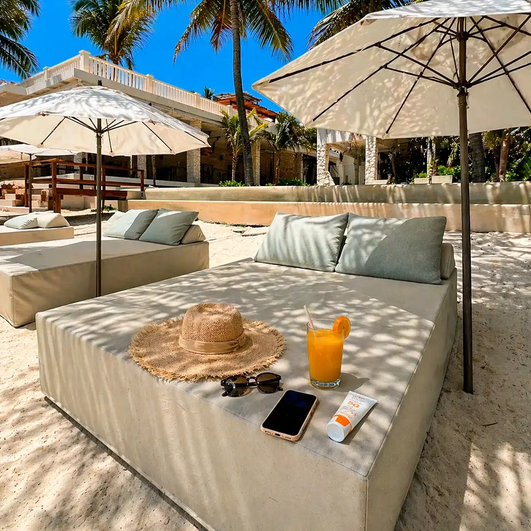 Cama de playa con cojines bajo una sombrilla, con un sombrero de palma y una bebida encima.