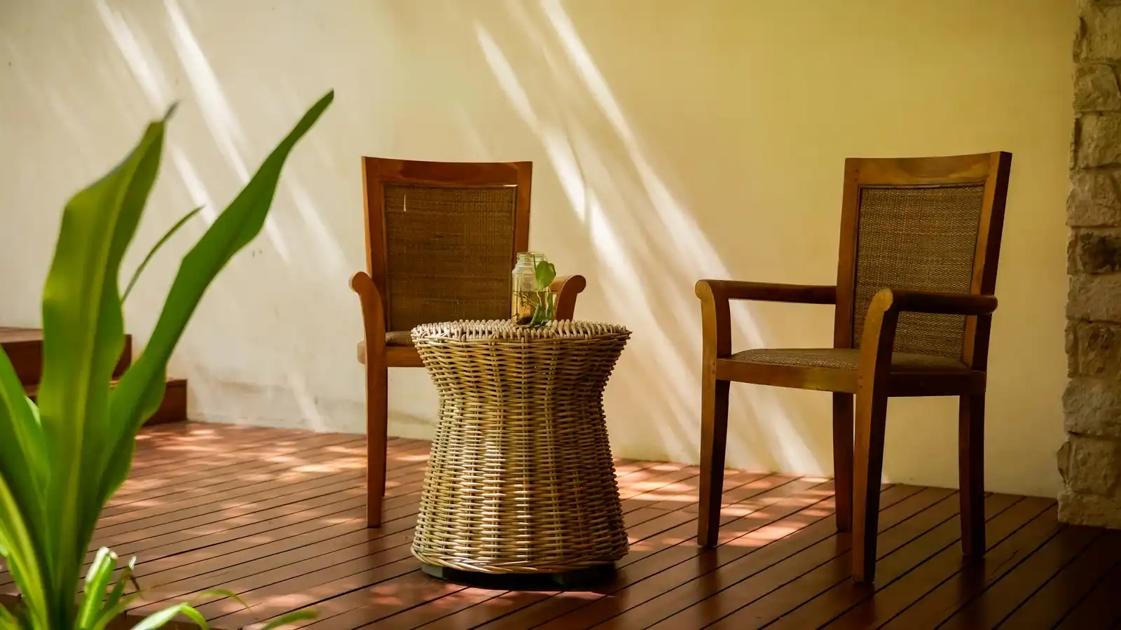 Two wooden chairs and a wicker table on a deck, beside a pale wall.