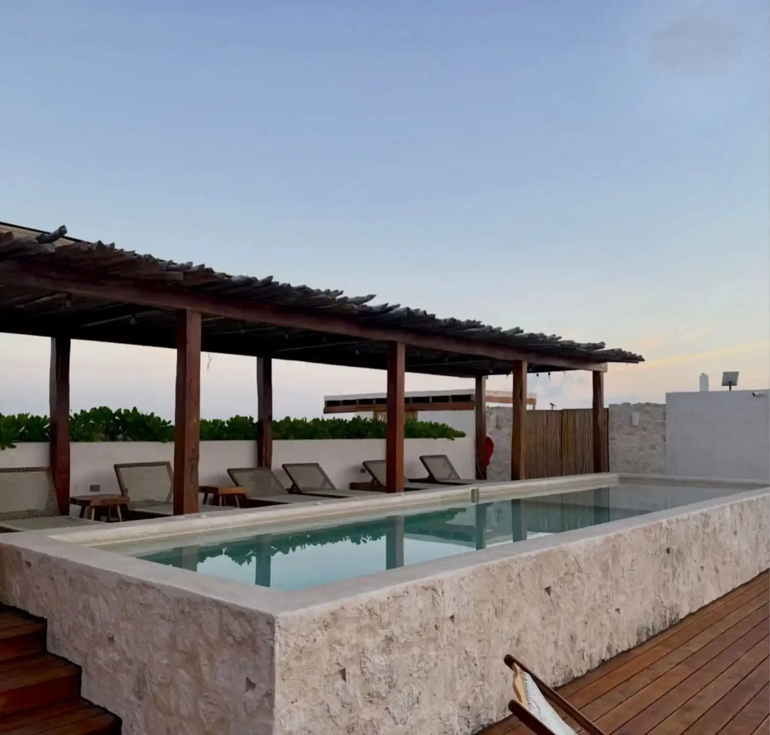 A stone-edged pool at dusk, under a wooden pergola, with a row of loungers and a hammock over the deck.