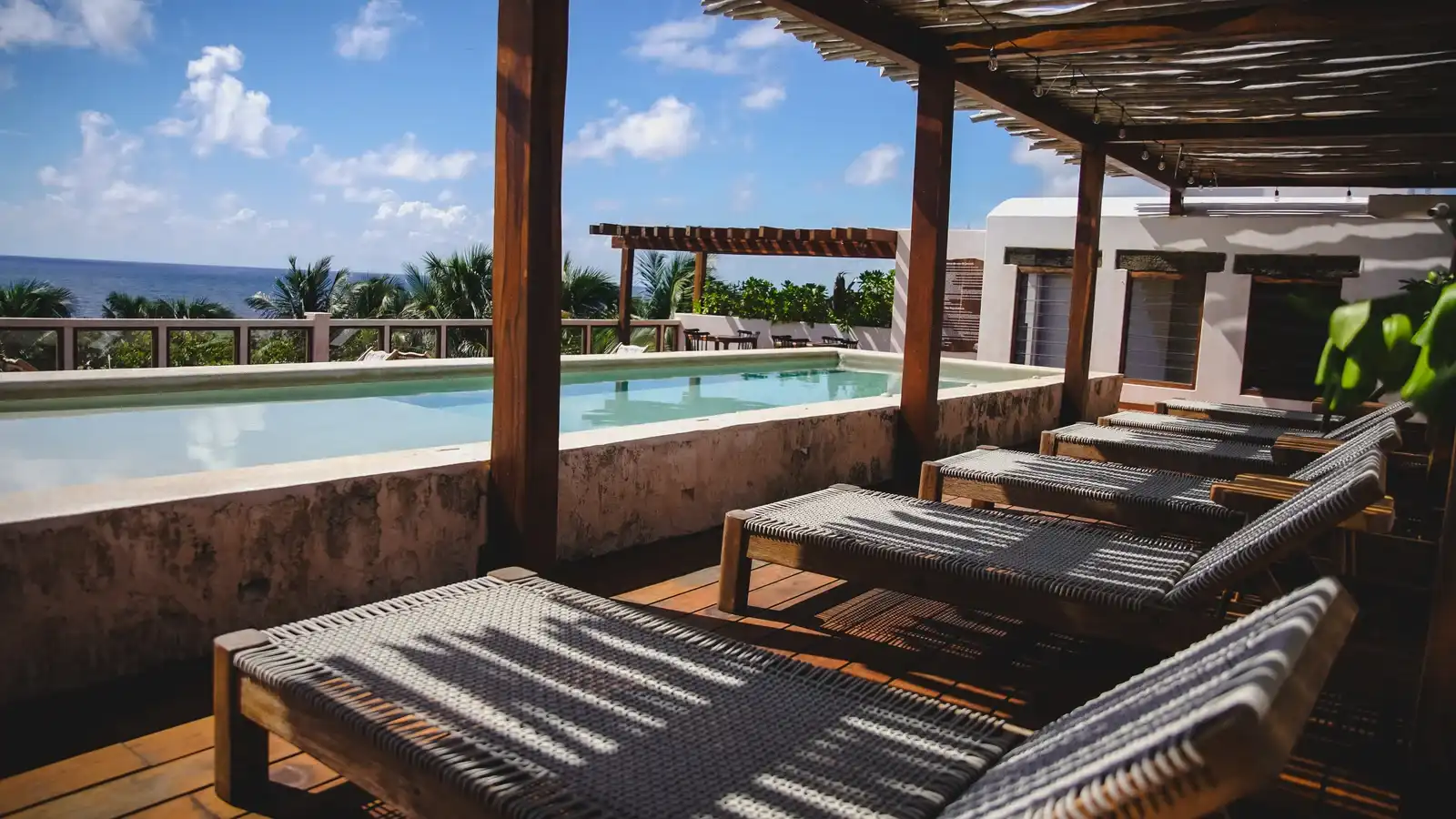 A long rooftop pool, with a wooden pergola, loungers and the sea beyond.