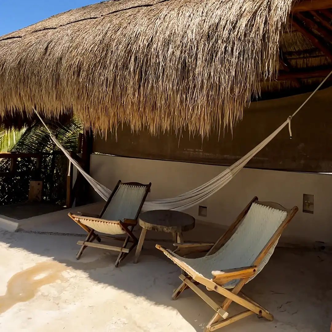 A palapa on the sand with a hammock and two wooden folding chairs.