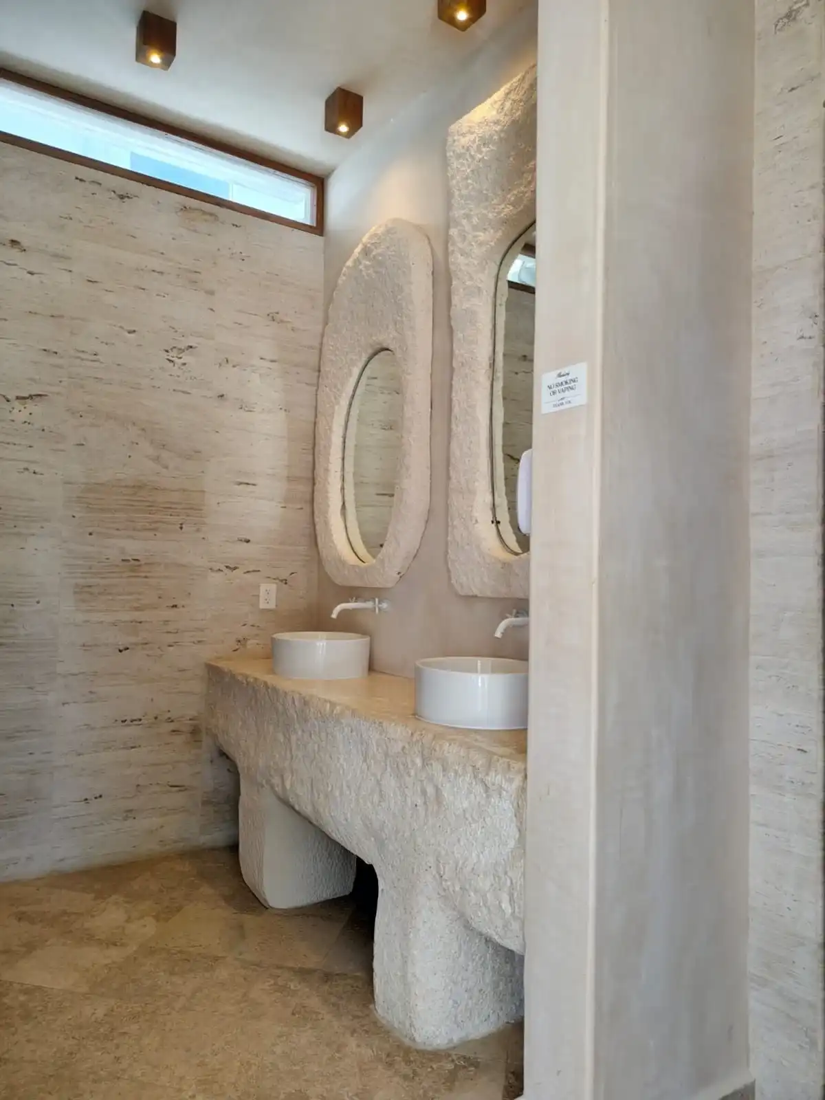 Travertine bathroom with two oval mirrors framed in stone and two round basins on a carved counter.