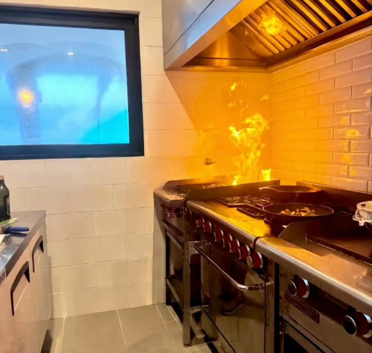 La cocina en marcha: una sartén al fuego sobre la parrilla, con la llama viva bajo la campana de acero.