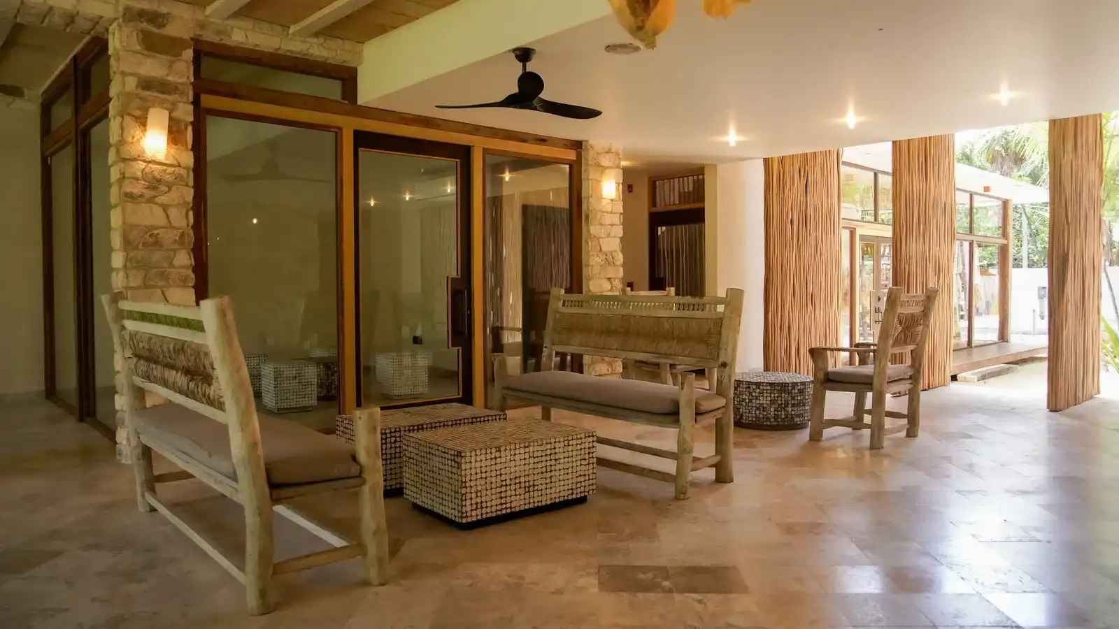 A sitting room with wooden armchairs, large windows and a ceiling fan.