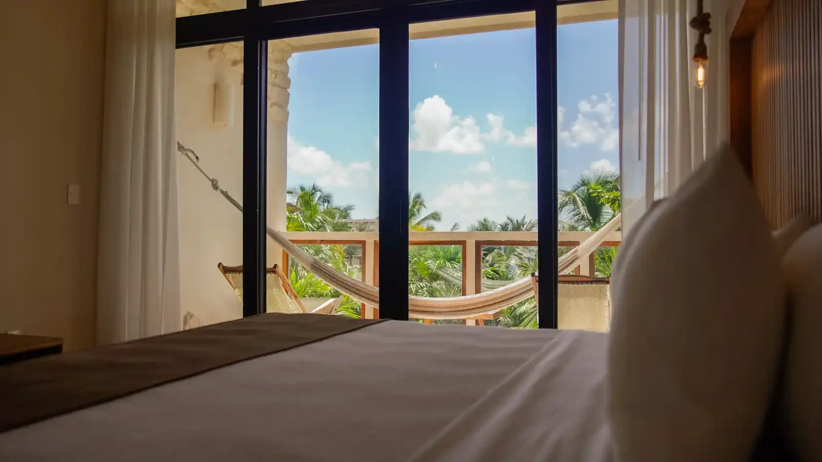 A bed facing a window wall, with a hammock on the balcony and the sea between the palms.