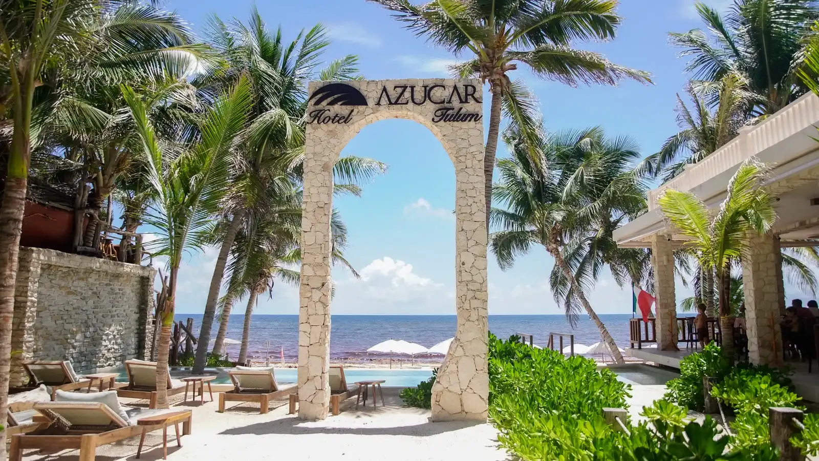 A stone arch bearing the hotel sign among palm trees, with the beach loungers and the Caribbean behind.